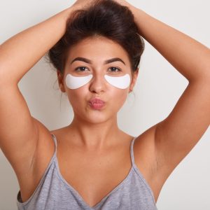 Studio shot of young beautiful woman with clean perfect skin with cosmetic patches on face. Beauty portrait of charming girl keeps hands up and lips rounded, looks at camera, isolated over white wall.